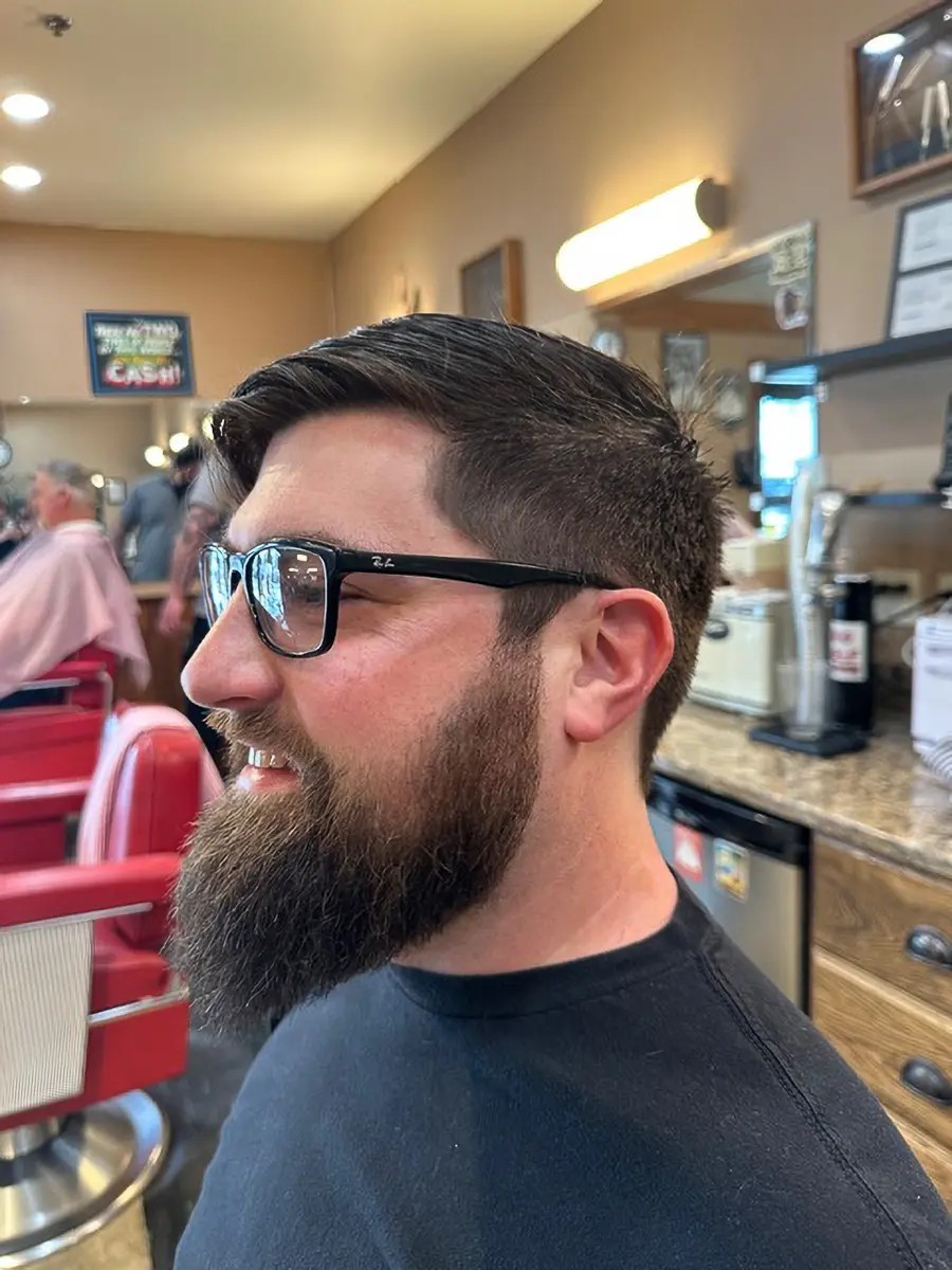 Side profile of a man with a neatly trimmed beard and short haircut wearing black-framed glasses inside a barbershop.