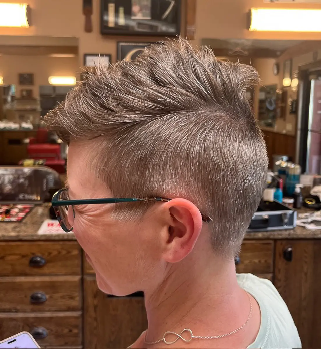Side view of a person with short, spiked gray hair wearing glasses and a silver infinity necklace inside a barber shop.