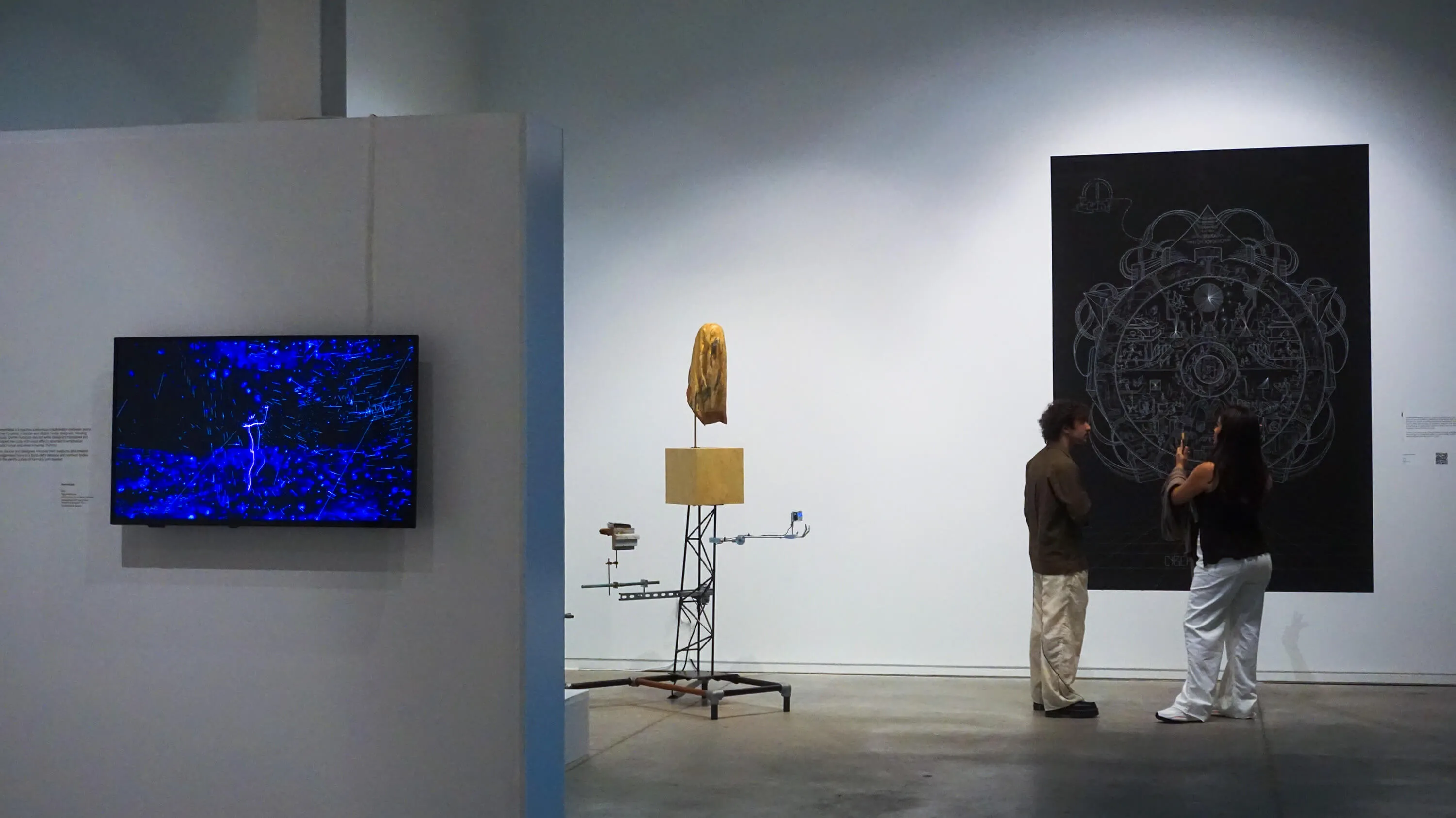 Two people standing in an art gallery observing a large circular intricate black and white artwork on the wall and a sculptural installation with a golden face atop a cube on metal framework.