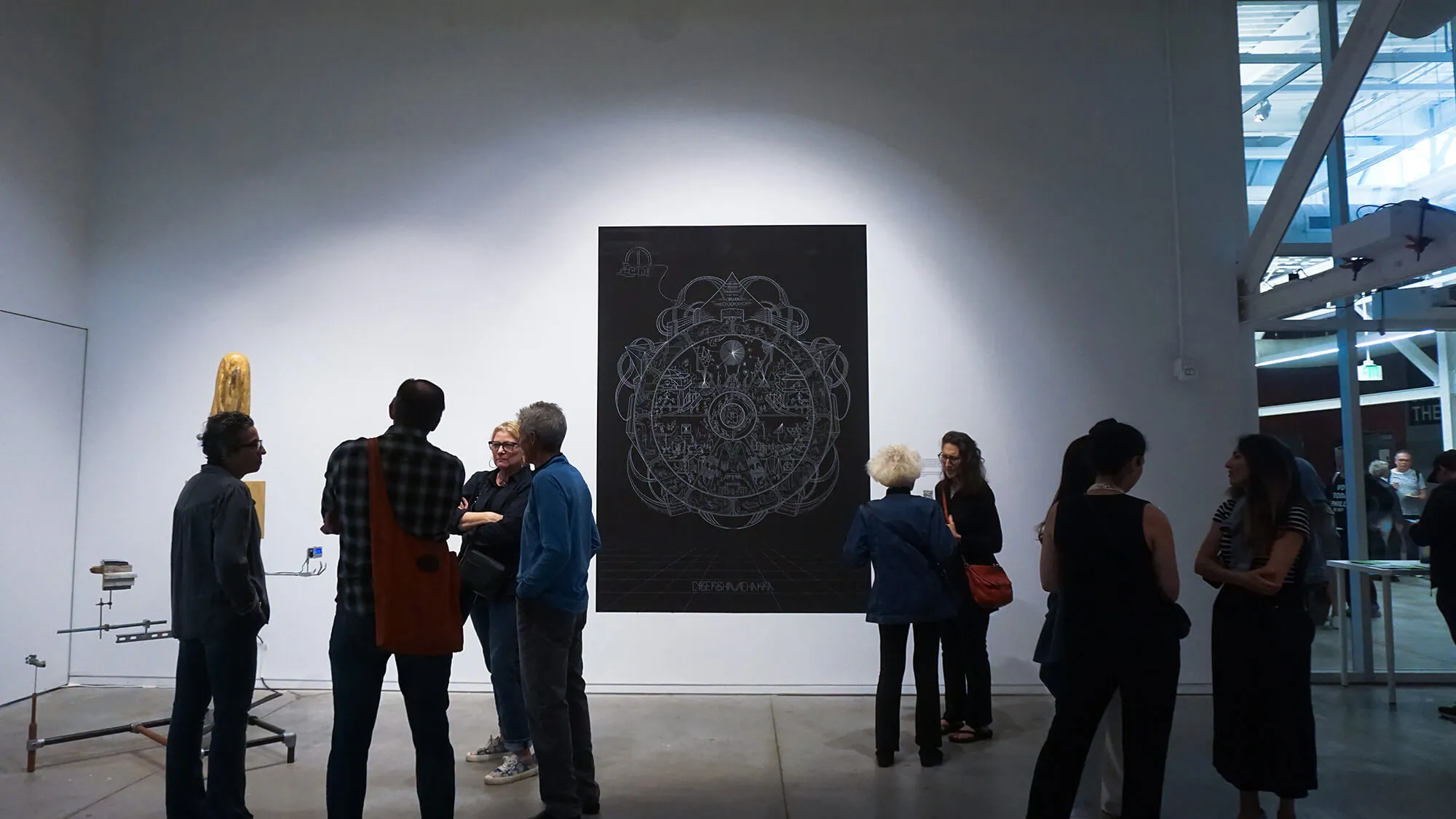 Group of people observing and discussing art in a gallery, with a large black and white detailed circular artwork on the wall.