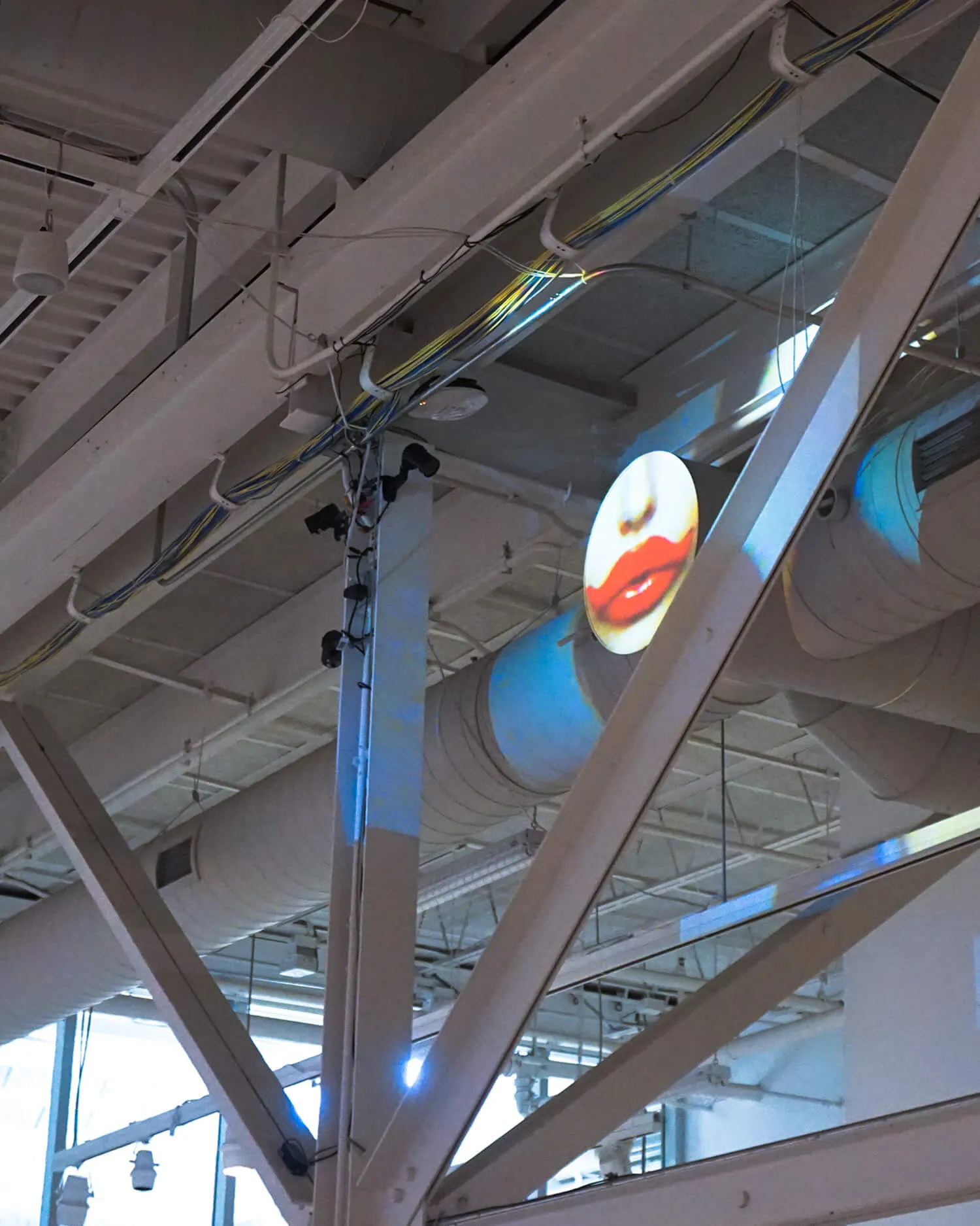 Ceiling with industrial beams, ductwork, cables, and a circular projection showing painted red lips.