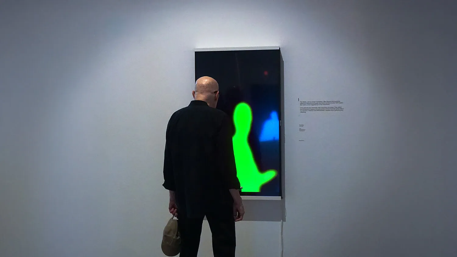 Bald man in black clothes facing and observing a digital artwork with green and blue abstract human silhouettes on a screen mounted on a white wall.