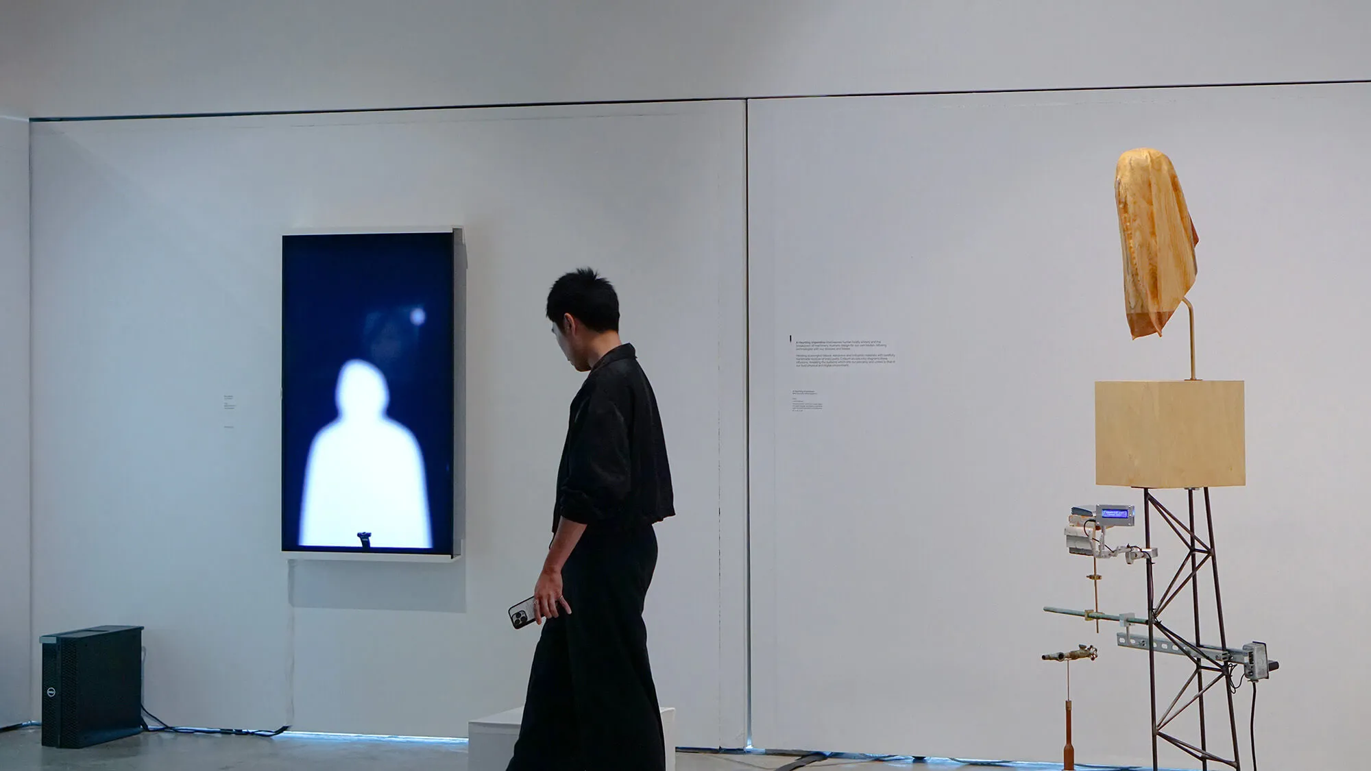 Man in black outfit holding a smartphone stands in an art gallery with a digital screen showing a blurry white silhouette and a wooden sculpture on metal framework.