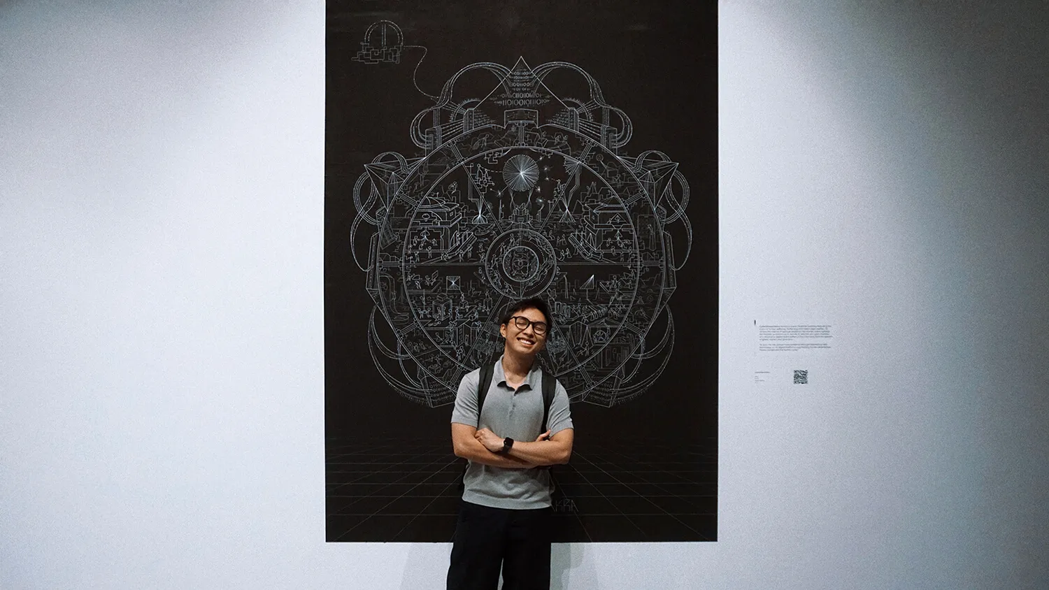 Smiling me with backpack stands in front of the Cyberbhavachakra poster featuring a complex, circular geometric design on a white gallery wall.