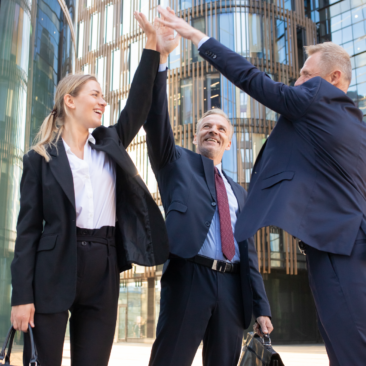 Three business professionals in suits giving a high-five in front of tall glass buildings.