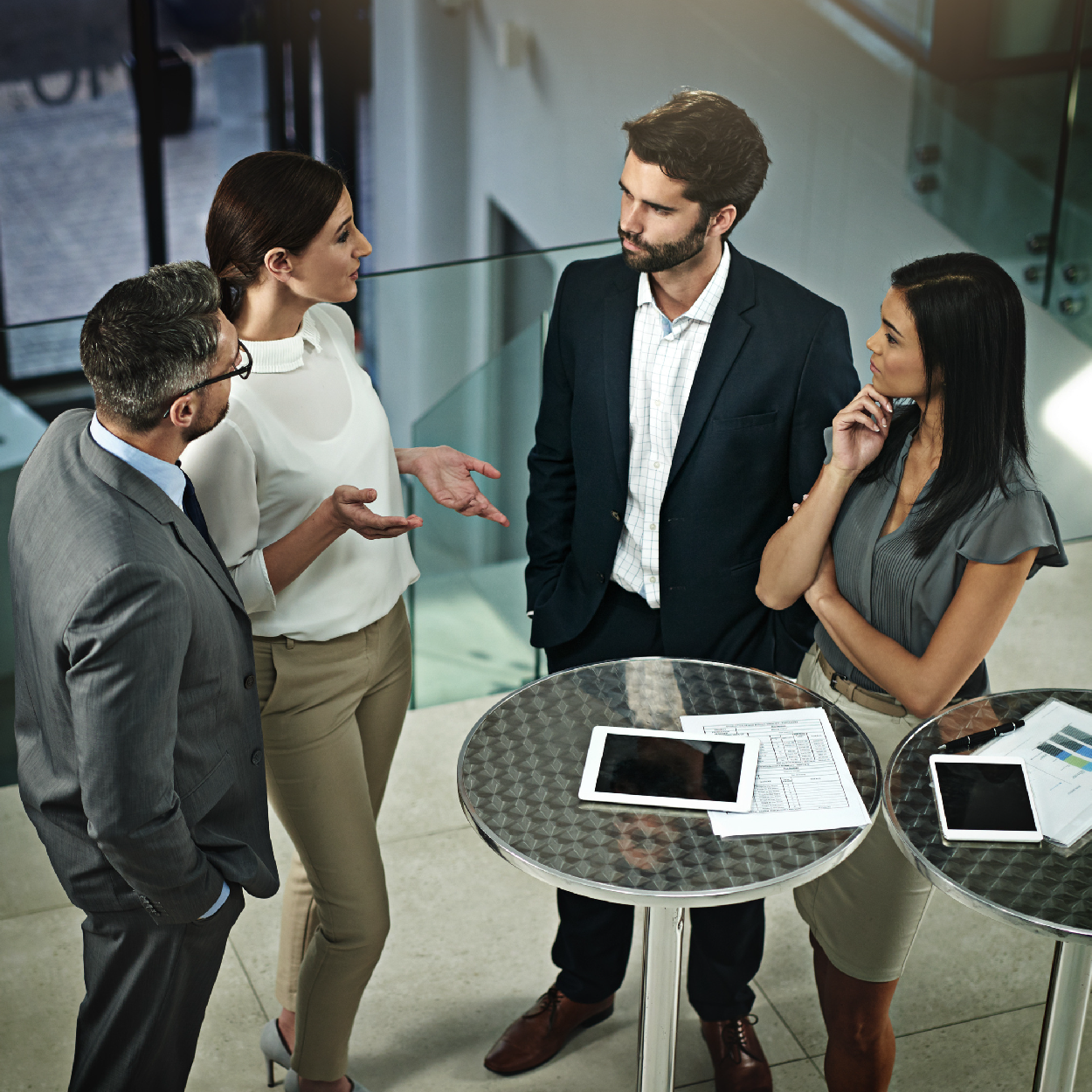 Four professionals having a serious discussion around high tables with tablets and documents in a modern office setting.