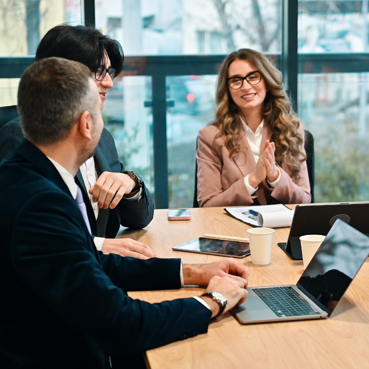 Three business professionals having a meeting and discussion around a table with laptops, tablets, and coffee cups.