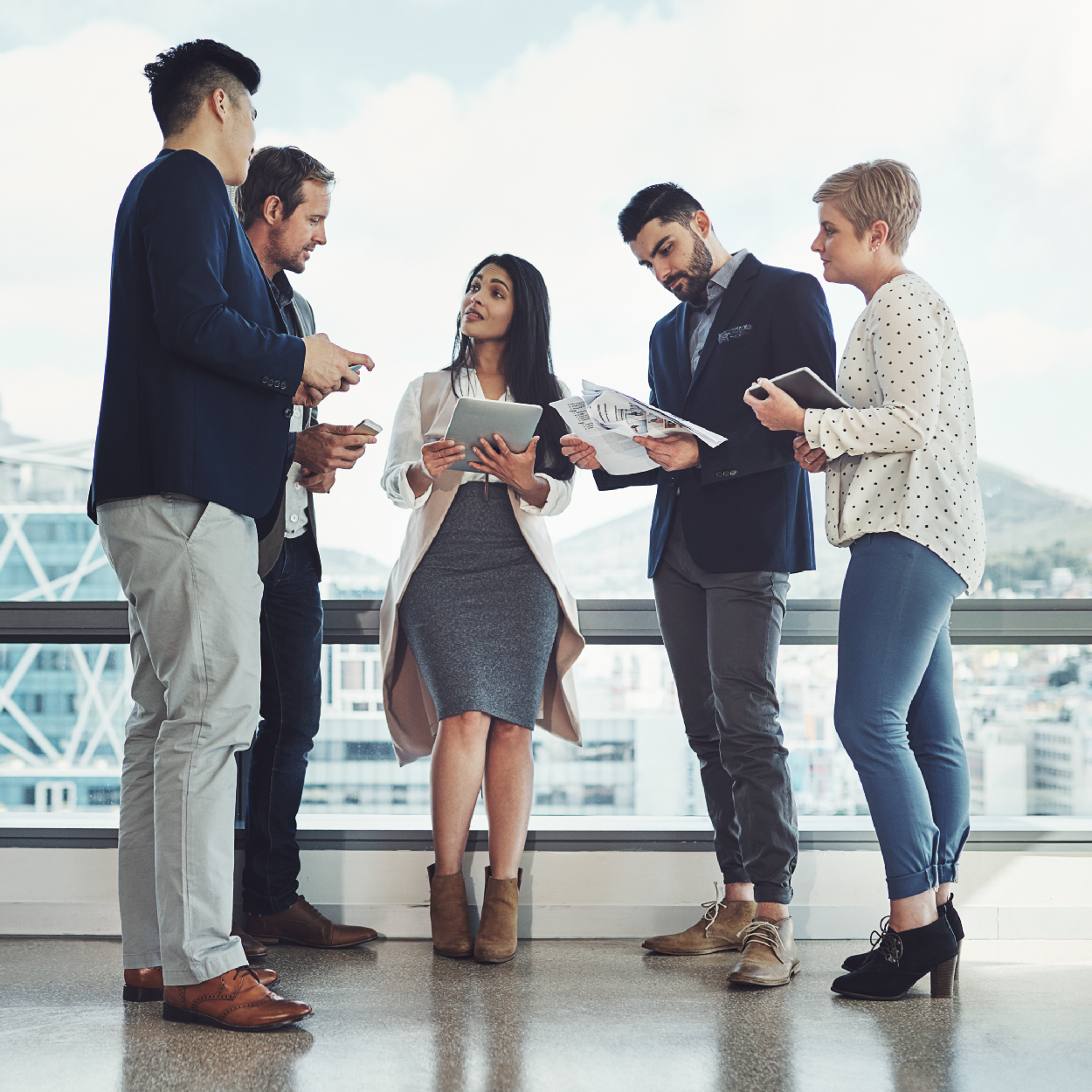 Five diverse professionals standing in a modern office discussing while holding tablets and documents.