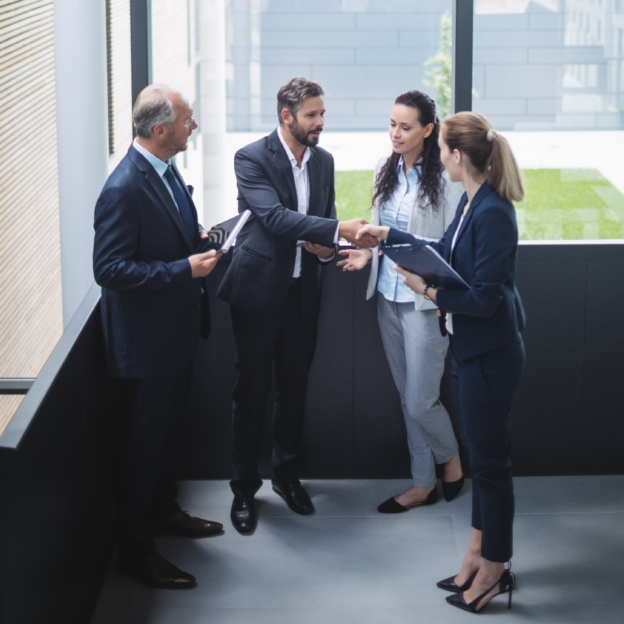 Two businesswomen and two businessmen in a bright office corridor, one man shaking hands with a woman holding a clipboard.