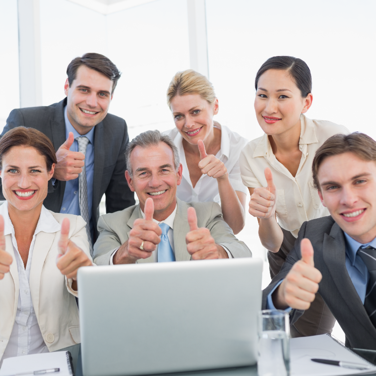 A group of six diverse businesspeople smiling and giving thumbs up around a laptop in an office.