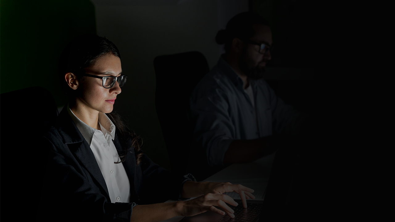 Focused woman wearing glasses and a blazer typing on a laptop in a dimly lit office with a man working in the background.