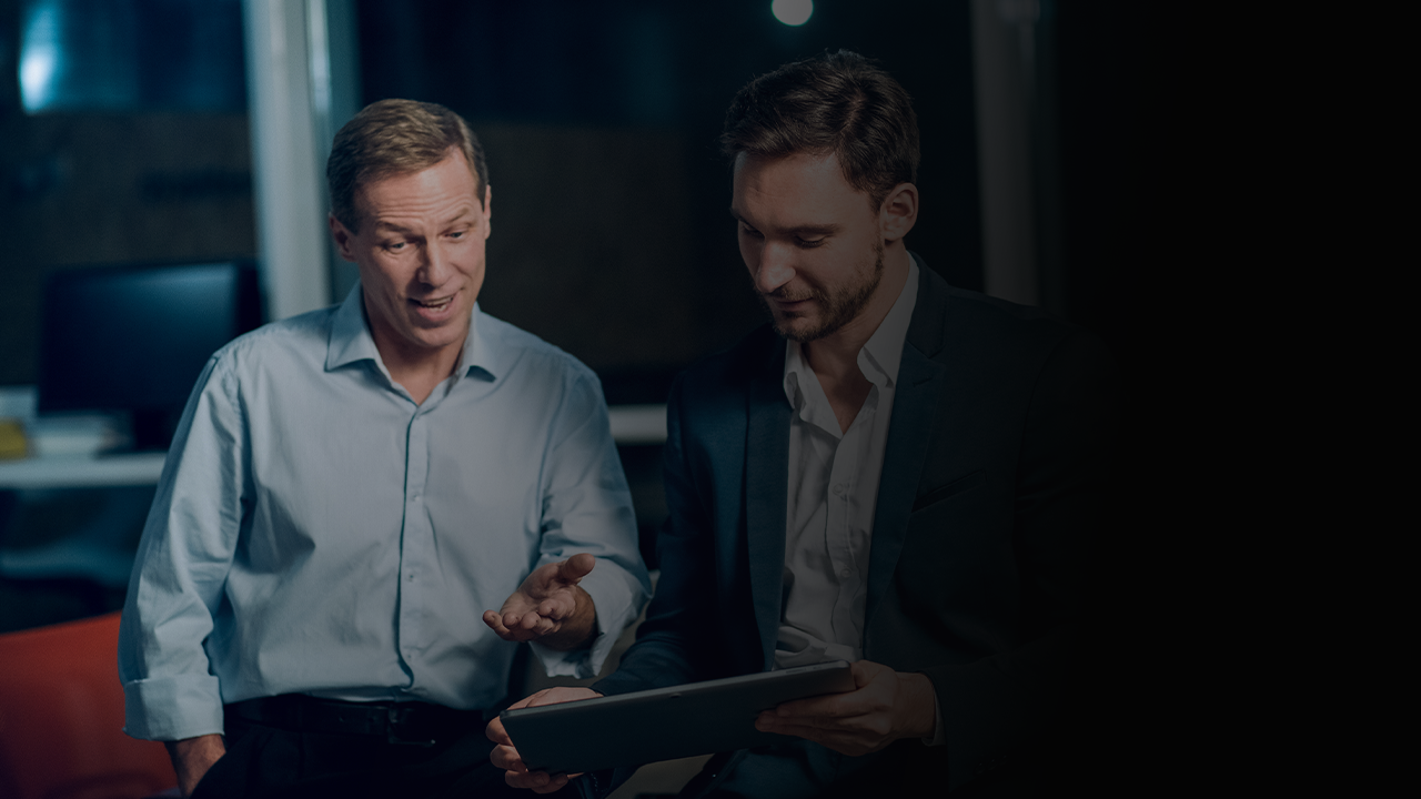Two men in business attire looking at a tablet during a discussion in an office setting.