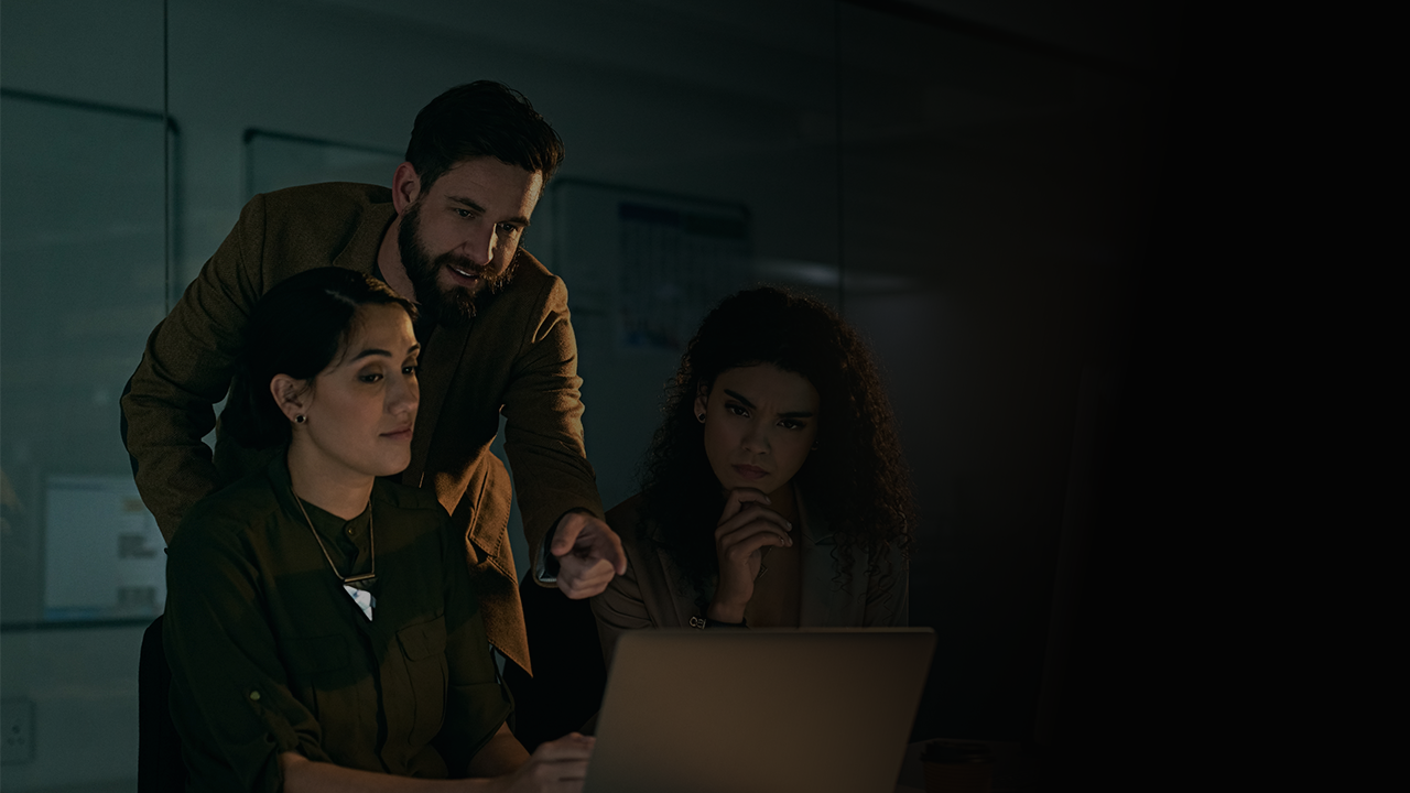Three colleagues in a dimly lit office looking intently at a laptop screen, one man pointing at the screen.