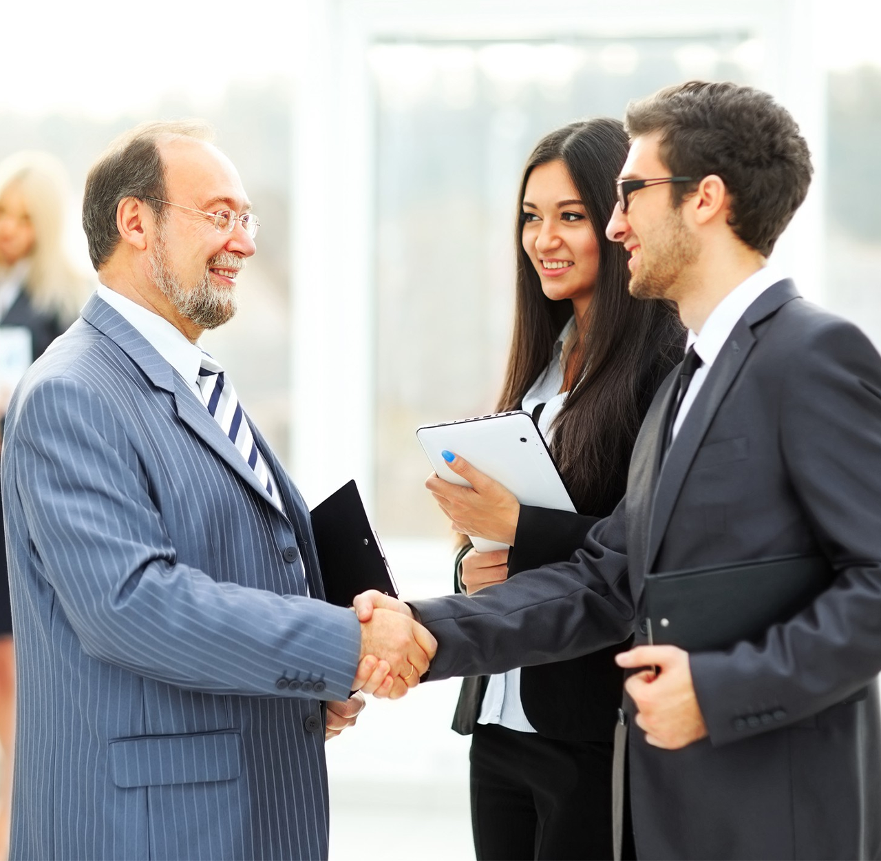 Two businessmen shaking hands while a businesswoman stands beside them holding a tablet, all smiling in a bright office setting.