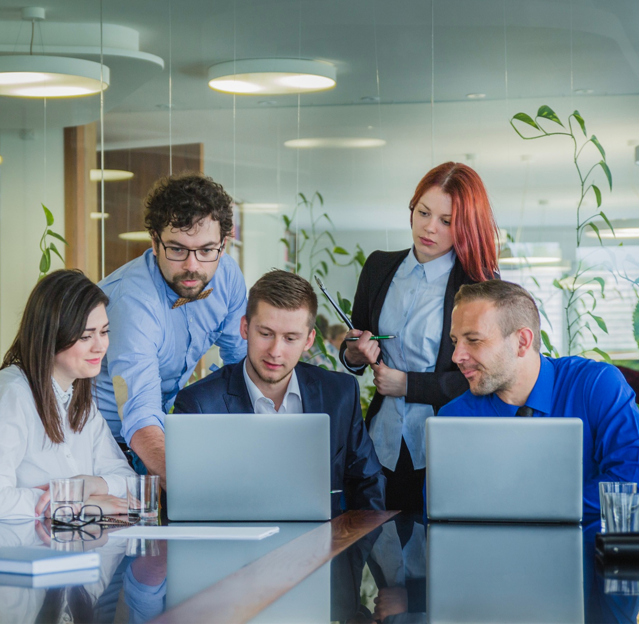 Five business professionals collaborating and looking at laptops in a modern office conference room.