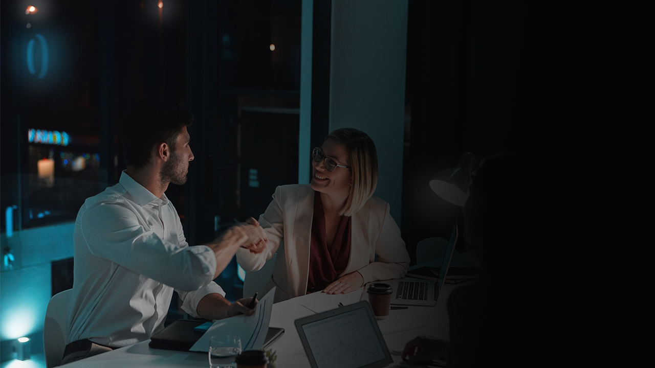 Two business professionals shaking hands across a table in a dimly lit office.