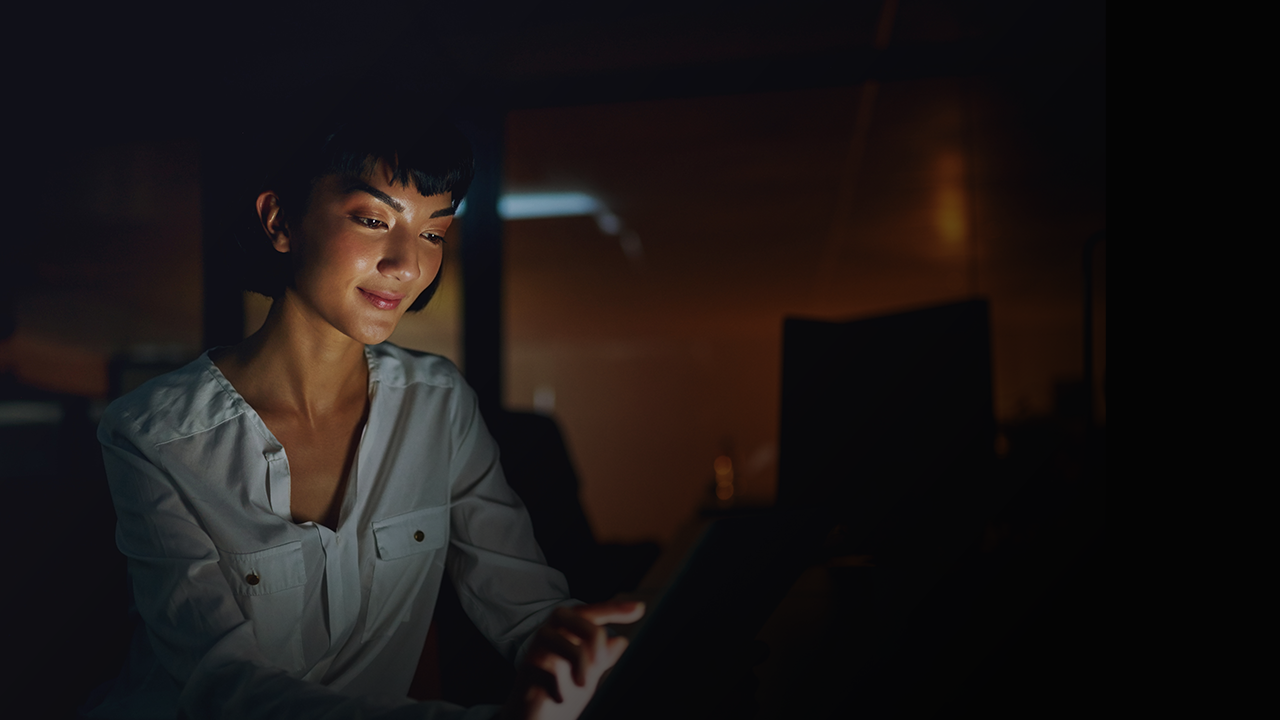 Woman in a white shirt smiling while using a tablet in a dimly lit room.