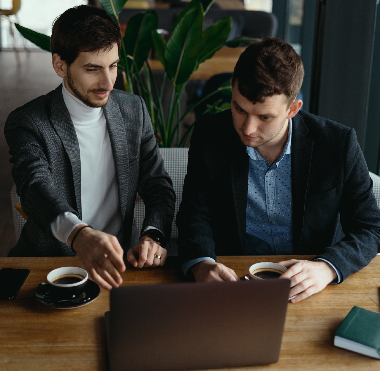 Two men in business attire sitting at a table with coffee cups, looking at and pointing to a laptop screen.