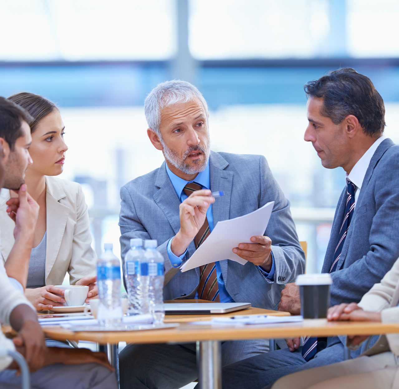Business meeting with four professionals in suits discussing documents around a table with water bottles and coffee cups.