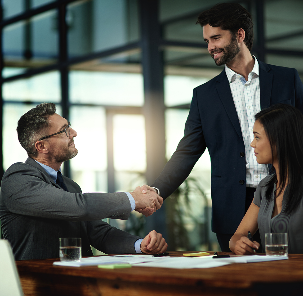 Two businessmen shaking hands across a table in a modern office while a woman looks on.