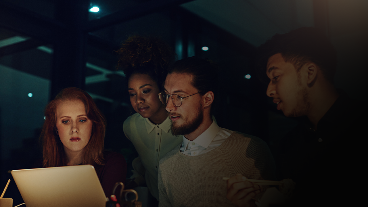 Four diverse young adults gathered closely, focused on a laptop screen in a dimly lit room.