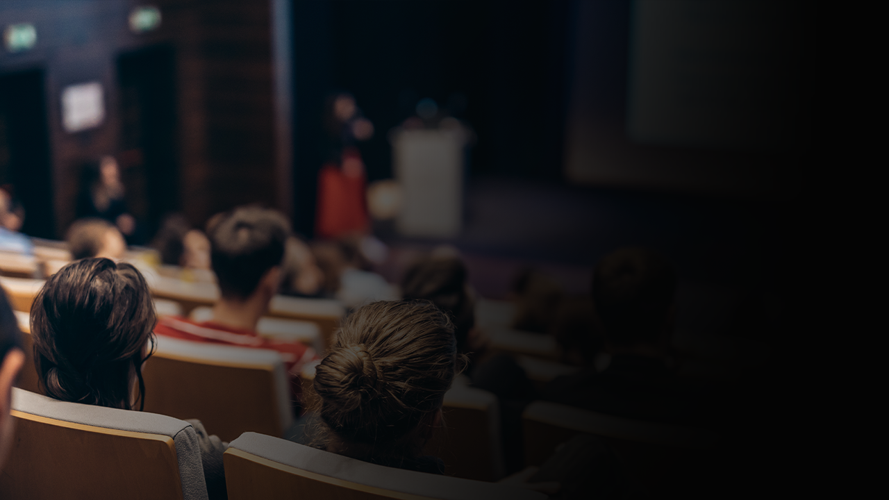 Audience seated in a lecture hall listening to a speaker at a podium.
