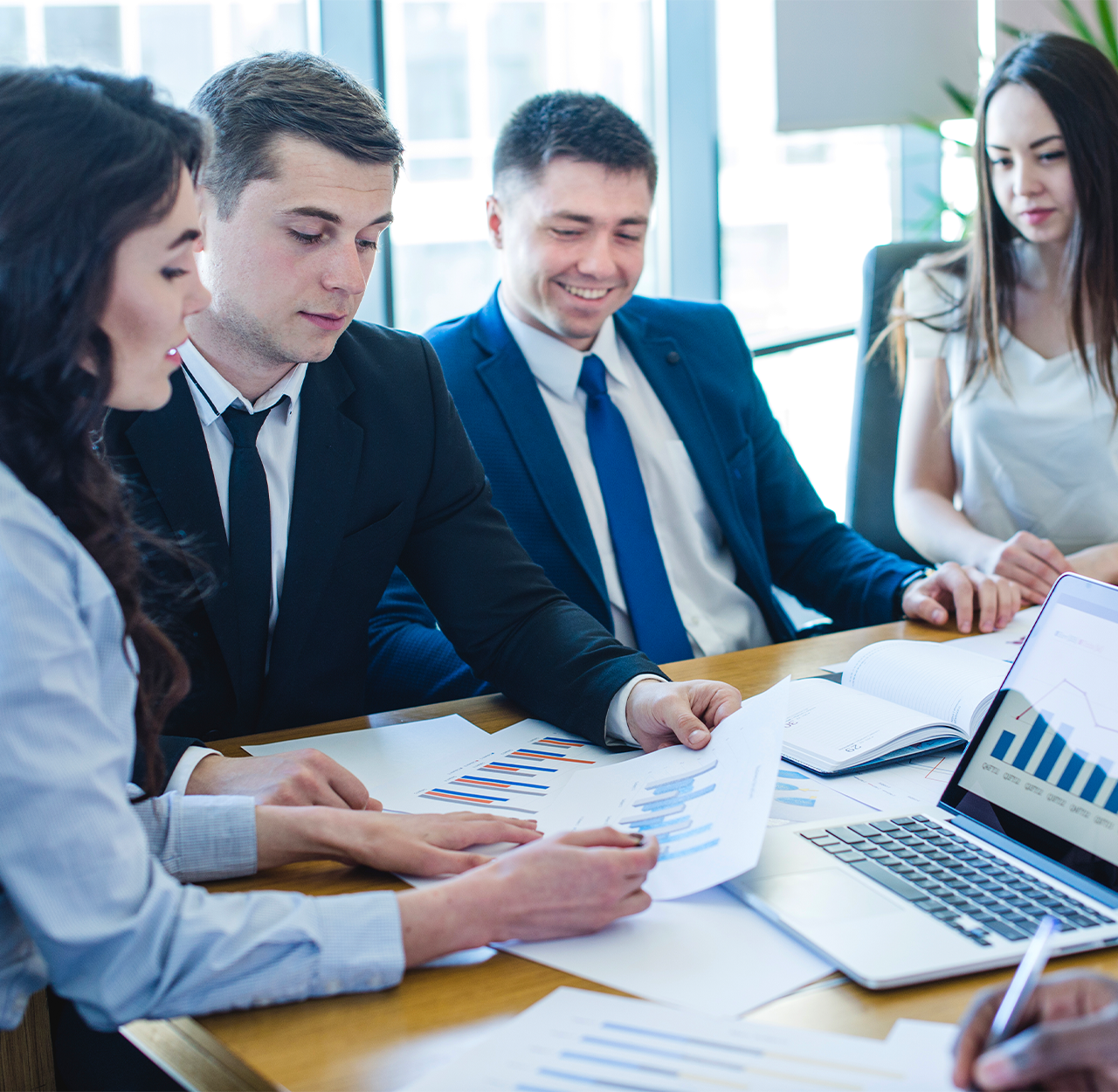 Four business professionals reviewing charts and graphs during a meeting around a conference table with a laptop displaying a bar chart.