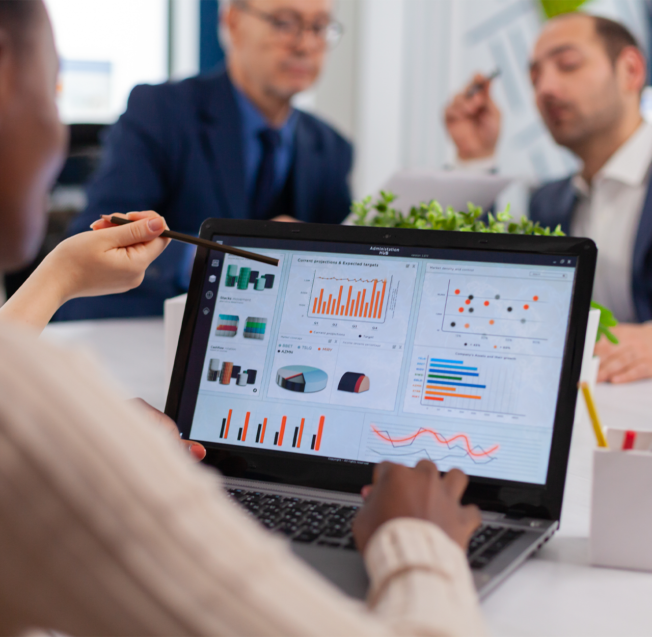 Person pointing at financial charts and graphs on a laptop screen during a business meeting with two blurred colleagues in the background.