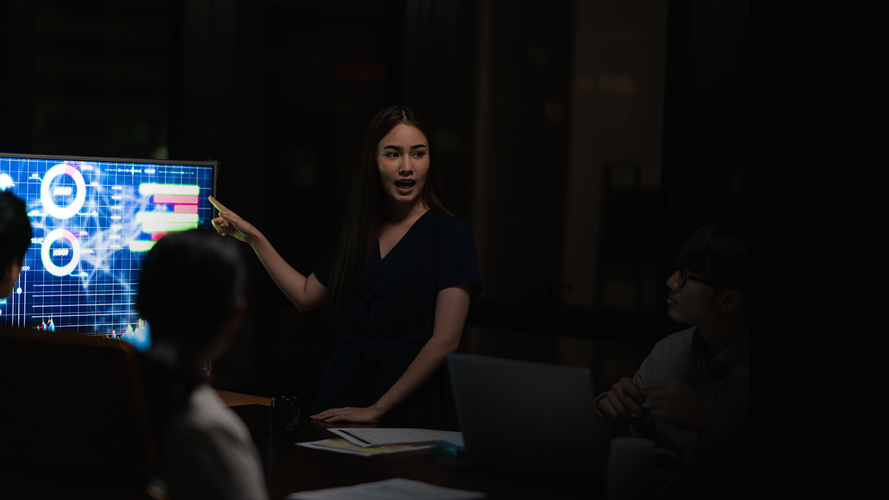 Woman presenting data on a screen with charts and graphs to colleagues in a dark meeting room.