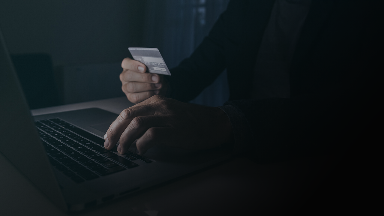 Person using laptop while holding a credit card for online payment in a dimly lit room.