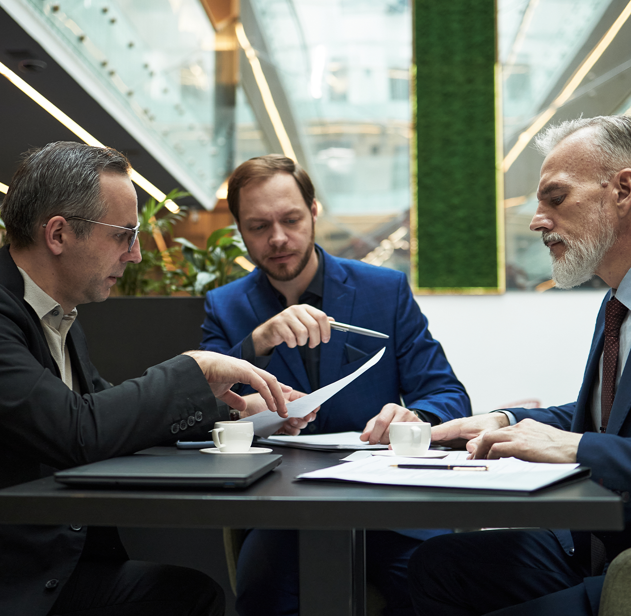 Three businessmen in suits having a discussion over documents at a modern office table with coffee cups.