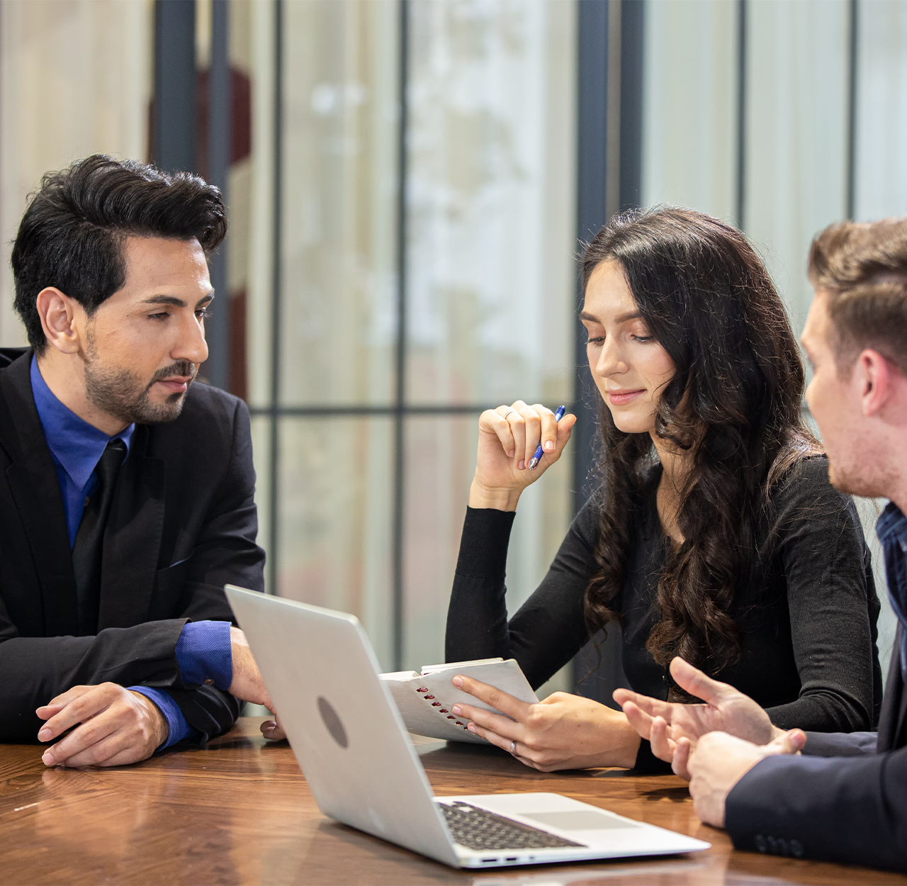 Three business professionals in a meeting reviewing notes and a laptop at a wooden table.