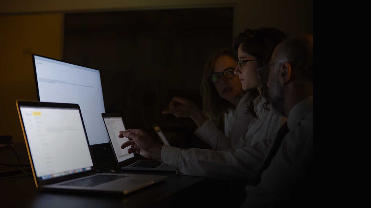 Three people working together on laptops and a monitor in a dimly lit room.