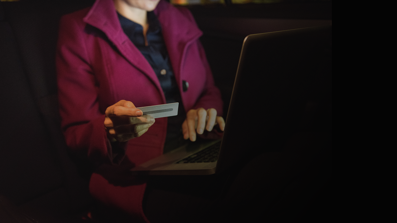 Person in dark clothing and red jacket holding a card and typing on a laptop in dim lighting.