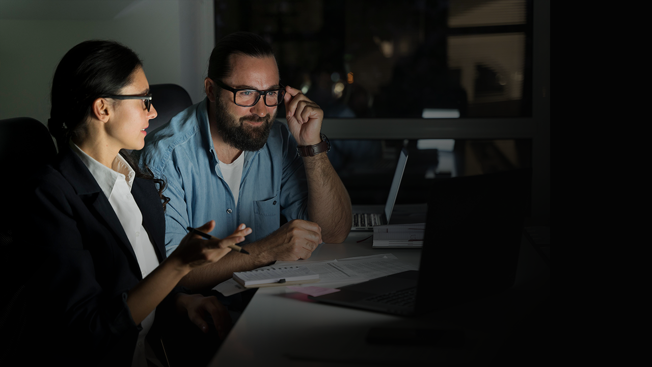 Two colleagues wearing glasses working together at a desk with laptops and documents in a dimly lit office.