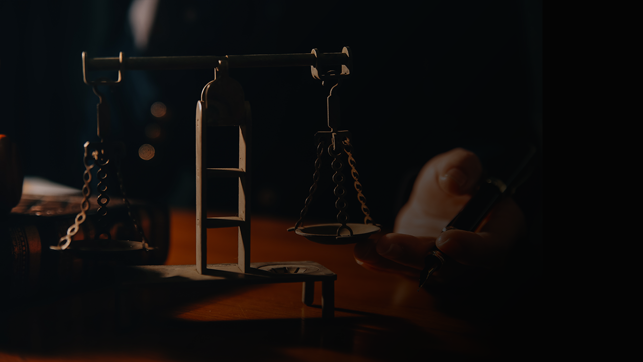 Close-up of a miniature balance scale on a wooden desk with a hand holding a pen in the background.