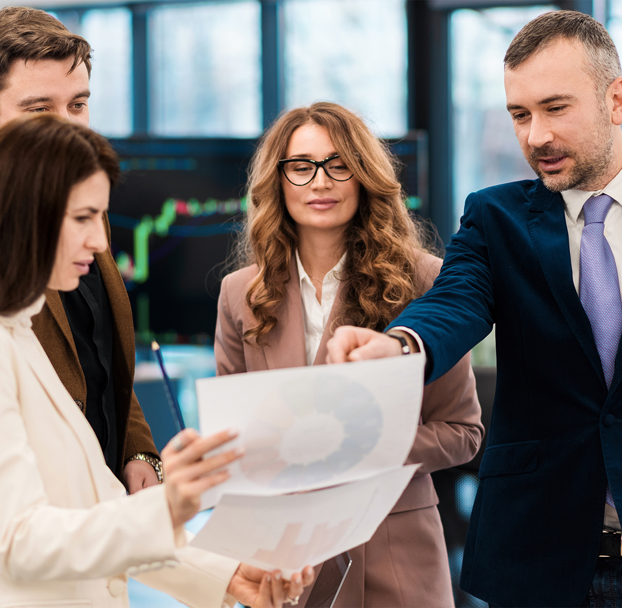 Four business professionals review and discuss charts in a modern office with large windows.