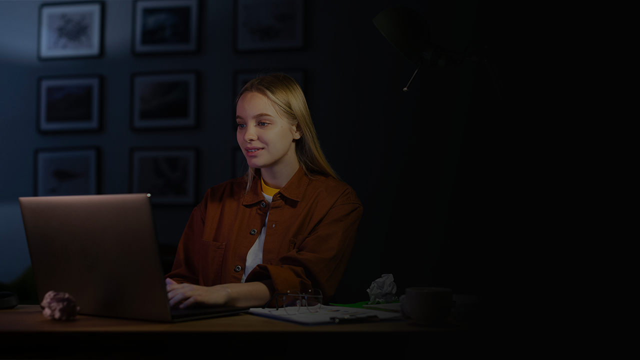 Young woman with long blonde hair working on a laptop at a desk in a dimly lit room with framed pictures on the wall.