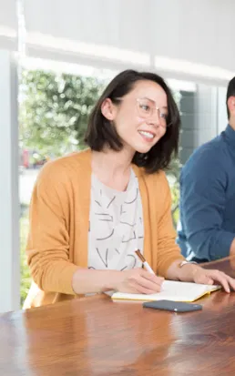 Femme souriante avec lunettes et cardigan orange prenant des notes à une table en bois dans un bureau lumineux.
