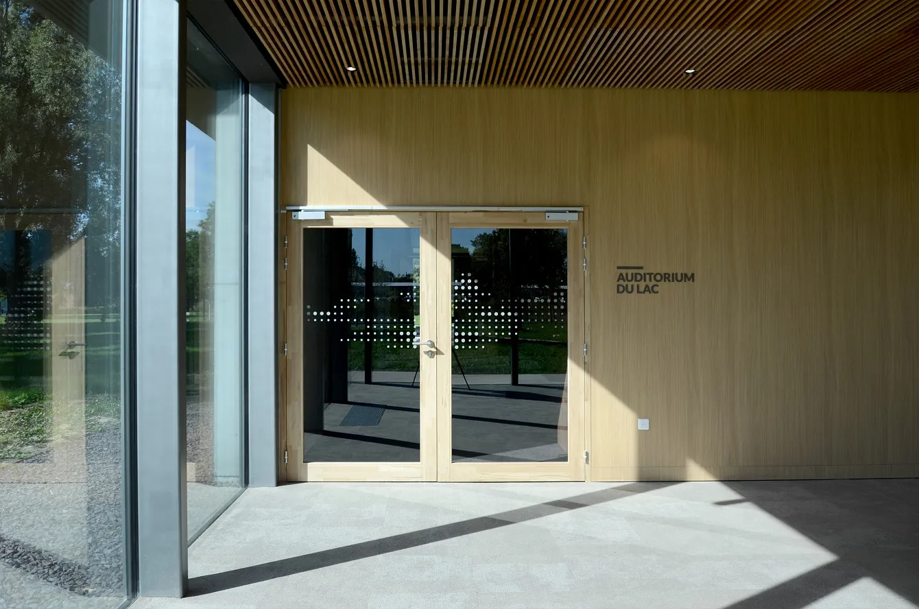 Entrée en bois avec double porte vitrée menant à l'Auditorium du Lac, murs en bois clair et grandes fenêtres sur le côté.