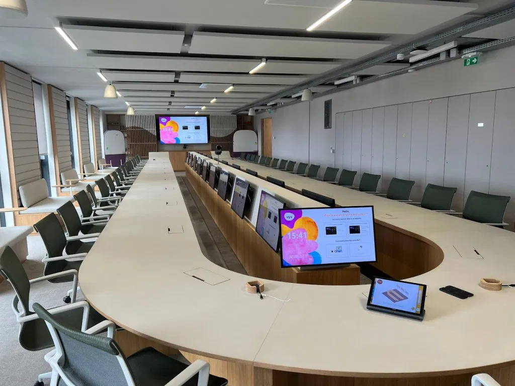 Grande salle de conférence moderne avec une table en forme de U équipée de nombreux écrans et chaises autour.