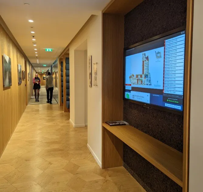 Couloir de bureau avec sol en parquet clair, des œuvres accrochées aux murs et un écran numérique intégré affichant un plan de bâtiment ; deux personnes discutent au fond du couloir.