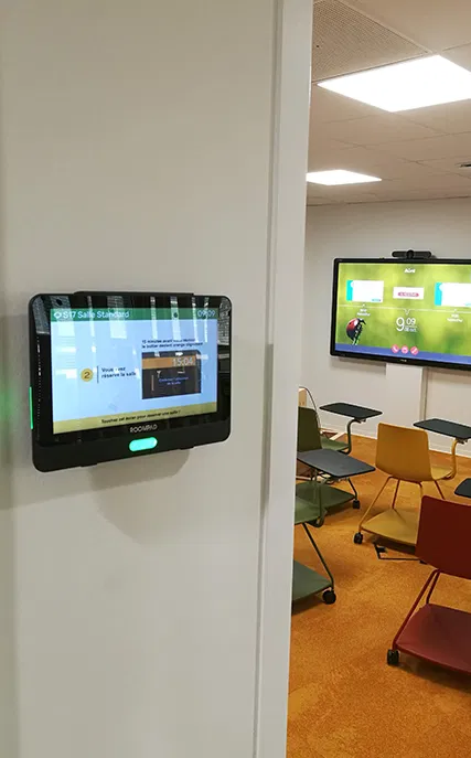 Salle de réunion avec des chaises de différentes couleurs autour de bureaux mobiles et deux écrans numériques, l'un fixé sur un mur et l'autre sur une cloison.