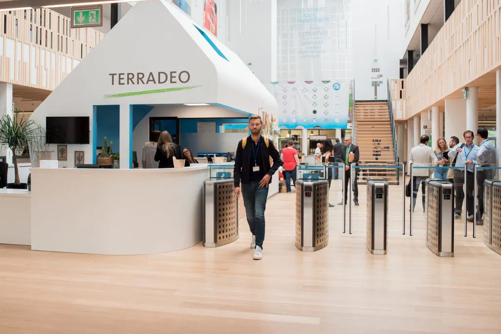 Un homme marche devant un comptoir d'accueil moderne portant le logo TERRADEO dans un grand hall lumineux avec plusieurs personnes en arrière-plan.