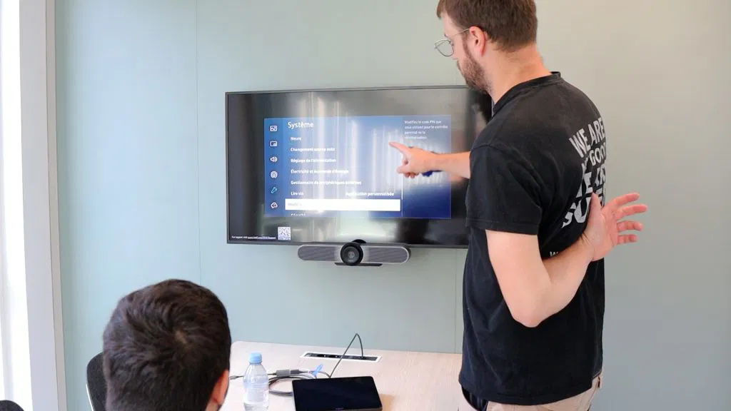 Un homme pointant l'écran d'une télévision murale affichant des paramètres système devant une autre personne assise à une table avec une tablette et une bouteille d'eau.