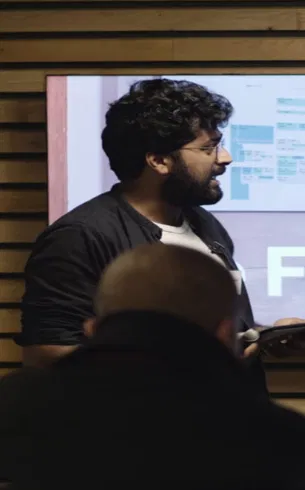 Un homme barbu aux cheveux bouclés portant des lunettes parle devant un écran avec un diagramme, à côté d'une personne de dos.