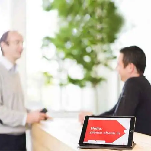 Deux hommes discutent au comptoir d'accueil avec une tablette affichant un message en anglais demandant de s'enregistrer.