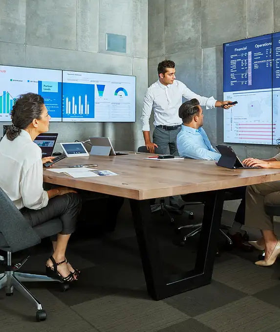 Quatre professionnels en réunion dans une salle de conférence moderne, avec des écrans affichant des graphiques et des données financières.