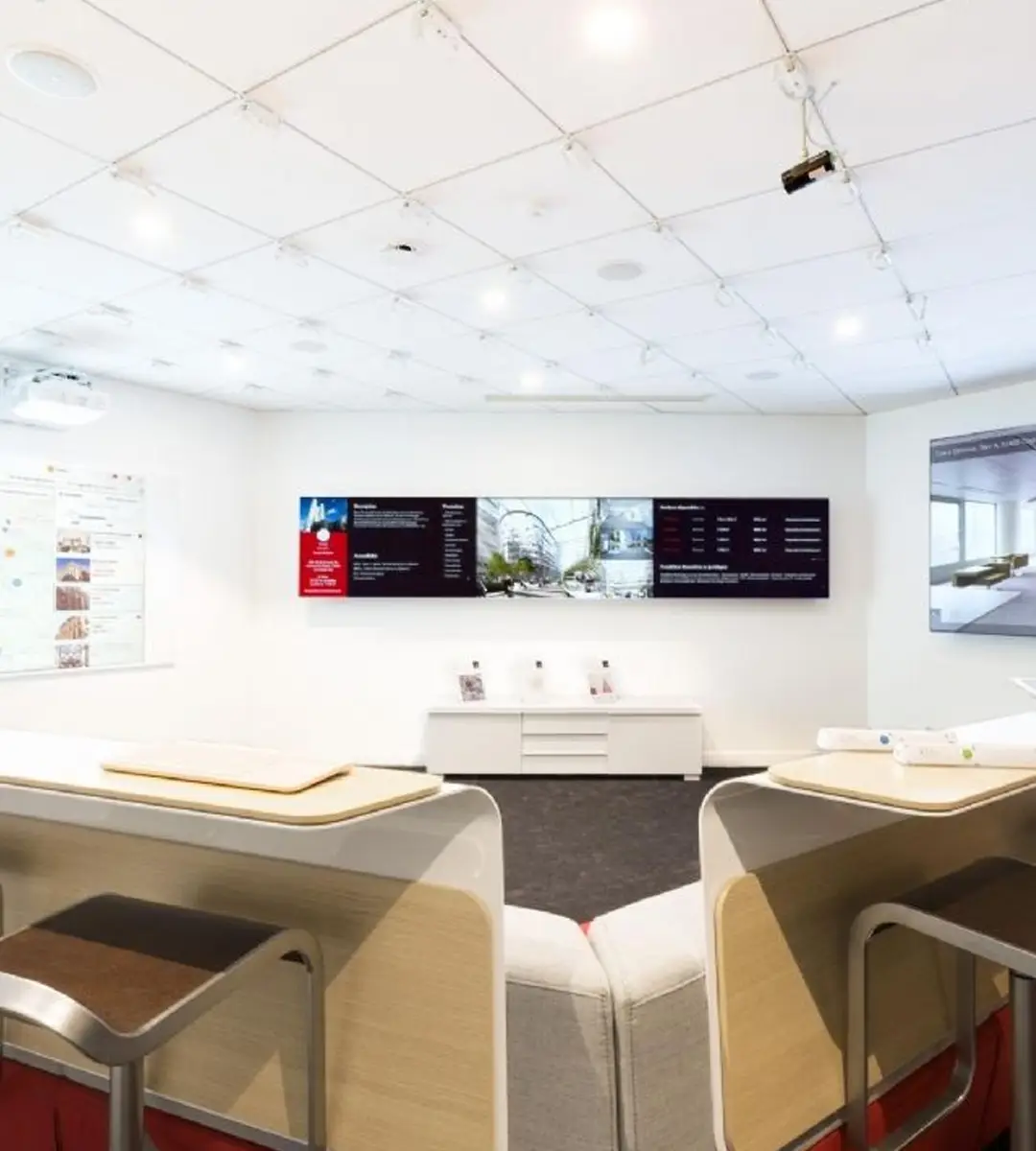 Salle lumineuse avec fauteuils et tables, un grand écran mural affichant des informations et une fenêtre laissant entrer la lumière naturelle.
