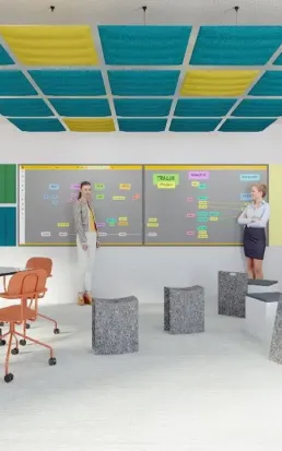 Deux femmes discutent devant un grand tableau noir couvert de notes colorées dans une salle de réunion moderne avec des sièges et un plafond décoré de panneaux carrés jaunes et bleus.